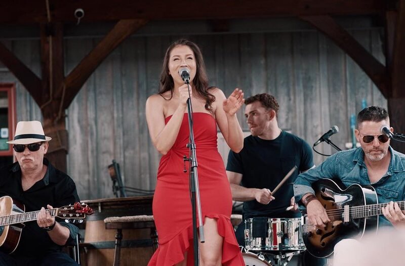 Woman in red dress singing into a microphone. Two men are sitting beside her, playing guitars. A man is behind her playing the drums.