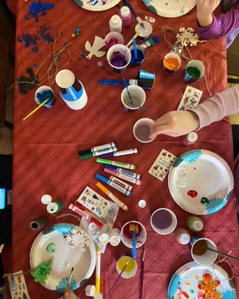 Arts and crafts spread across a table with a red tablecloth. 2 separate hands are seen reaching across the table to paint.
