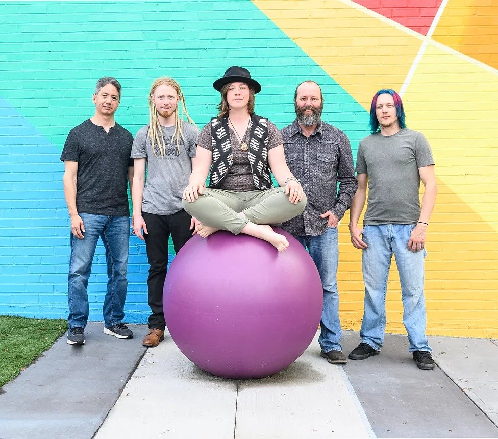 5 band members standing in front of colorful concrete wall. Middle band member is sitting on a purple ball.