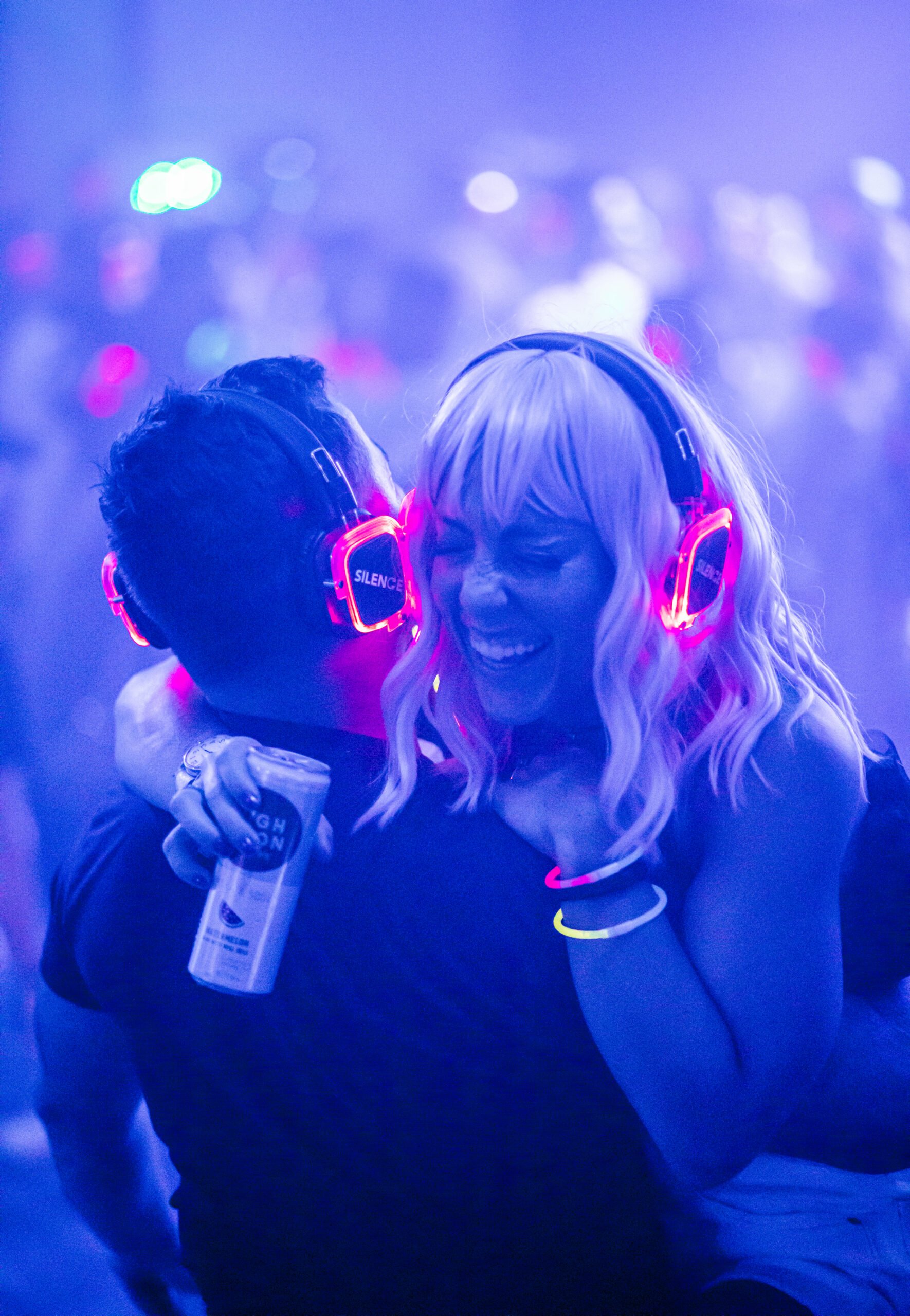 Man and woman dancing, wearing headphones. Woman is holding a drink and laughing.