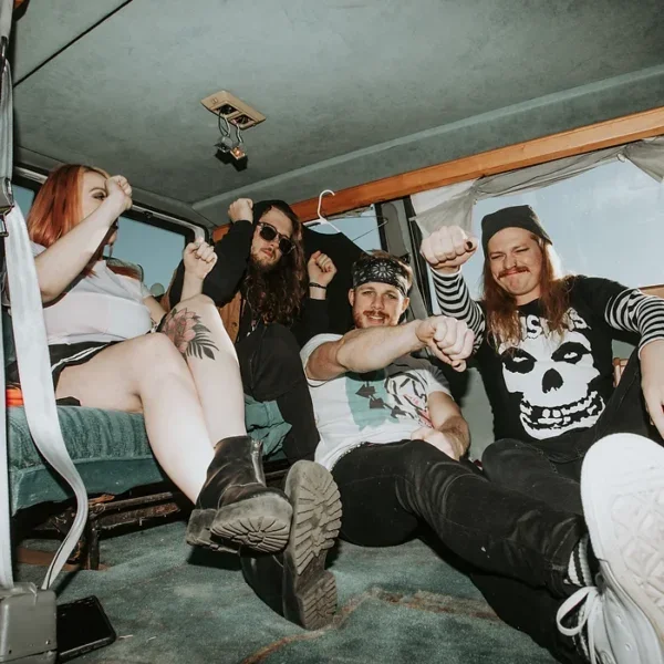 Five band members sit in a van dancing. Three men one women.