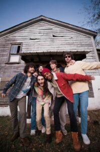 Six band members stand in front of barn house