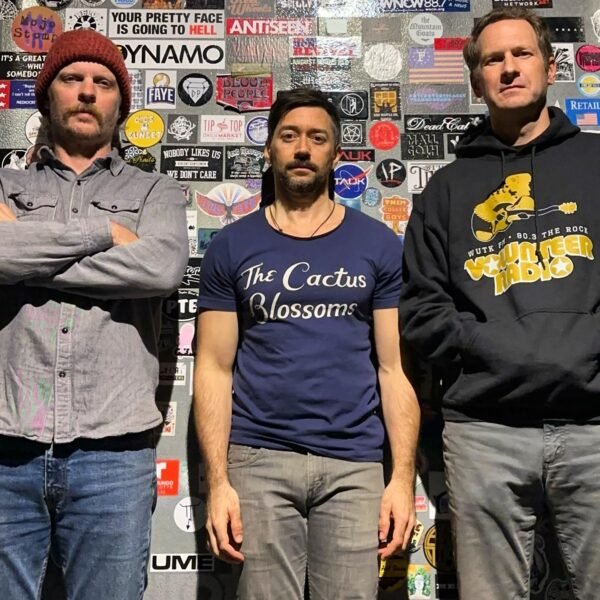 three men standing against a wall of stickers, casually dressed