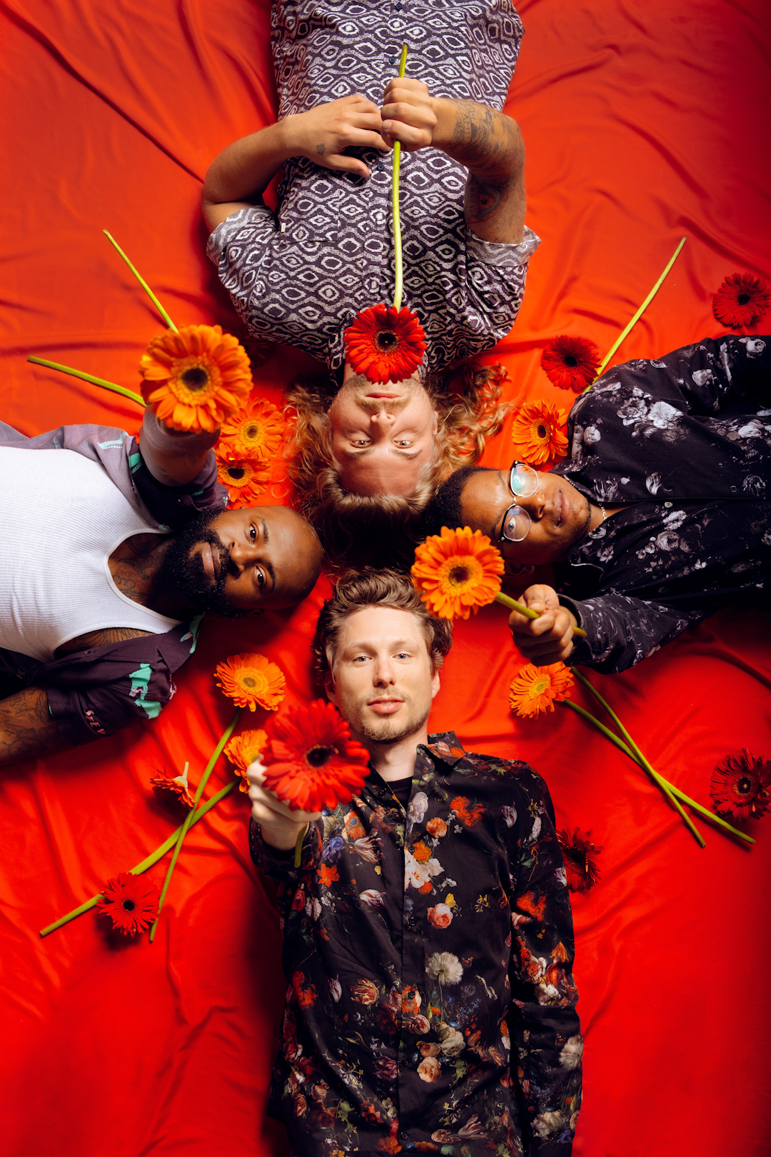 Four men laying down in a circle with an orange background surrounded by orange flowers